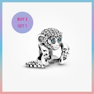 Puede incluir: Un charm de plata con forma de mono, con pequeñas piedras transparentes en la cabeza y las orejas. El mono tiene ojos azules y levanta los brazos. El texto "BUY 2 GET 1" está en un círculo morado.