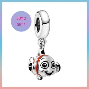 May include: A silver charm featuring a cartoon clownfish design with black and red accents. The charm has a large eye and a smiling face. The charm is suspended from a silver loop. A purple circle with the text "BUY 2 GET 1" is in the upper left corner.