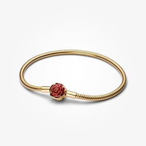 Puede incluir: Pulsera dorada con un charm de rosa roja. La pulsera tiene un diseño de cadena de serpiente y un cierre de broche. El charm de rosa está detallado con esmalte rojo y detalles dorados. La pulsera se muestra sobre un fondo blanco.