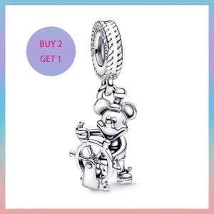 Puede incluir: Un charm de plata con Mickey Mouse sosteniendo un timón. El charm tiene una parte superior en forma de neumático con un diseño circular. La imagen incluye un círculo morado con el texto "BUY 2 GET 1".