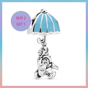Op de afbeelding: Een zilveren bedel met een stripfiguur die een blauw-witte paraplu vasthoudt. De bedel heeft een gedetailleerd ontwerp en hangt aan de paraplu. Een paarse cirkel met de tekst "BUY 2 GET 1" staat op de achtergrond.