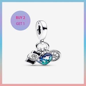 May include: A silver charm with travel-themed pendants. The charm features a camera with a clear stone, a heart-shaped world map, and a compass. The text "BUY 2 GET 1" is in a purple circle in the upper left corner.