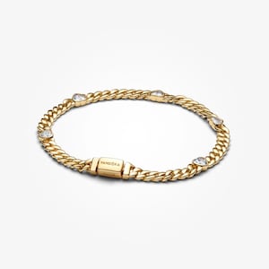 Op de afbeelding: Een goudkleurige armband met een gourmette kettingontwerp. De armband heeft hartvormige, heldere edelstenen en een sluiting met de merknaam "Pandora" gegraveerd. De sieraden worden op een witte achtergrond gepresenteerd.