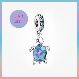 Può includere: Un charm in argento con un design a forma di tartaruga marina. La tartaruga ha un corpo blu e turchese simile al vetro e piccole pinne argentate. La parte superiore del charm ha un anello con pietre blu e trasparenti. È incluso anche il testo "BUY 2 GET 1".