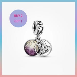 May include: A silver charm with a round pendant featuring a family of bears on a swing set. The charm also has a round pendant with a purple and white landscape design. The charm has the text "BUY 2 GET 1".