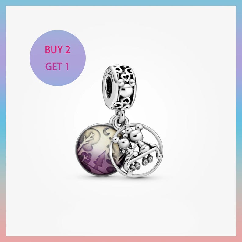 May include: A silver charm with a round pendant featuring a family of bears on a swing set. The charm also has a round pendant with a purple and white landscape design. The charm has the text "BUY 2 GET 1".