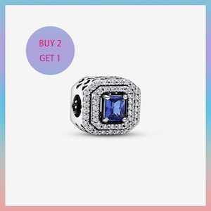 Peut inclure: Un charm en argent avec une pierre précieuse carrée bleu foncé au centre, entourée d'un double halo de pierres claires et scintillantes. Le charm présente des détails ajourés complexes sur les côtés. Le texte "BUY 2 GET 1" est dans un cercle violet.