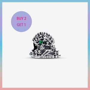 Puede incluir: Un charm de plata con un diseño arquitectónico detallado, con una pequeña gema verde. El charm tiene detalles intrincados y una superficie texturizada. Un círculo morado con el texto "BUY 2 GET 1" está en la esquina superior izquierda.