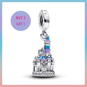 May include: A silver charm with a castle design, featuring blue, purple, and white enamel accents. The charm has a round top with a small gem and the text "PANDORA ALE S925". The charm is hanging from a silver ring. The image also includes a purple circle with the text "BUY 2 GET 1".