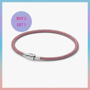 May include: A mauve-colored woven bracelet with a silver clasp. The bracelet is circular and appears to be made of a soft, flexible material. The image includes a purple circle with the text "BUY 2 GET 1".