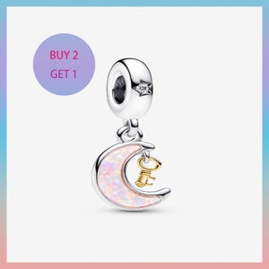 Puede incluir: Un charm de plata con un acento de estrella y una luna creciente colgante con una llave dorada. La luna tiene un diseño de ópalo rosa y blanco. El texto "BUY 2 GET 1" está en un círculo morado.