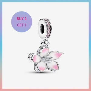 Op de afbeelding: Een zilveren bedel met een roze en witte bloemhanger. De bedel heeft een ring met roze edelstenen en de tekst "BUY 2 GET 1" in een paarse cirkel.