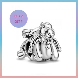 Può includere: Un charm d'argento a forma di cammello, con borse laterali dettagliate ed elementi decorativi. Il charm ha una faccia sorridente ed è posto su uno sfondo bianco. Un cerchio viola con la scritta "BUY 2 GET 1" si trova in alto a sinistra.