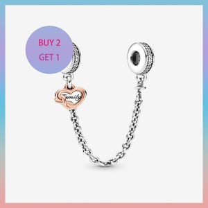 May include: A silver charm bracelet safety chain with a rose gold heart charm that says "Family". The charm is connected to two silver ends with clear stones. A purple circle with the text "BUY 2 GET 1" is in the background.
