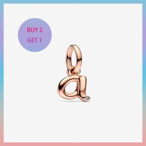 May include: Rose gold charm in the shape of the lowercase letter "a" with a small clear gemstone. The charm has a loop at the top for attaching to a bracelet or necklace. A purple circle with the text "BUY 2 GET 1" is in the background.