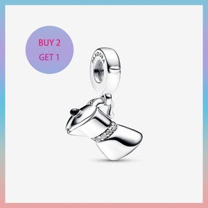 May include: A silver charm shaped like a coffee pot with a sparkling band of clear stones. The charm has a loop at the top for attaching to a bracelet or necklace. A purple circle with the text "BUY 2 GET 1" is in the background.