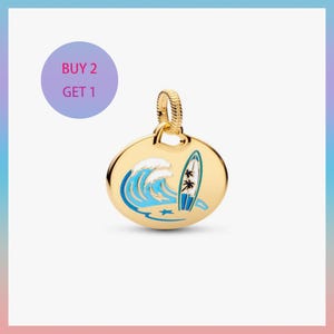 Op de afbeelding: Goudkleurige hanger met een strandthema. De hanger heeft een blauw-witte golf, een surfplank en twee palmbomen. De tekst "BUY 2 GET 1" staat in een paarse cirkel.