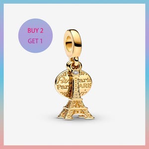 Puede incluir: Un charm dorado con la Torre Eiffel. El charm tiene un colgante redondo con la palabra "París" y una pequeña gema transparente. El charm está diseñado para ser adjuntado a una pulsera o collar.