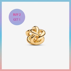 May include: A gold-colored charm with an intertwined knot design. The charm is a bright, shiny gold and has a smooth, polished surface. A purple circle with the text "BUY 2 GET 1" is in the upper left corner.