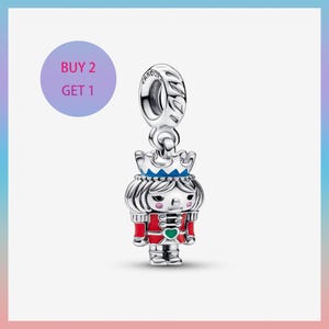 May include: A silver charm with a nutcracker design. The charm features a red and silver uniform, a blue crown, and a green heart. The charm is suspended from a silver loop. The text "BUY 2 GET 1" is in a purple circle.