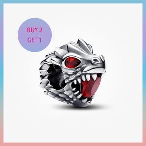 Puede incluir: Un charm de cabeza de dragón plateado con ojos de piedras preciosas rojas y una piedra preciosa roja en la boca. El charm tiene escamas detalladas y dientes afilados. Un círculo morado con el texto "BUY 2 GET 1" está en la esquina superior izquierda.