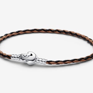 Puede incluir: Pulsera de cuero trenzado con cierre plateado. La pulsera presenta hebras de cuero negro y marrón. El cierre es de color plateado brillante. Esta pulsera es un accesorio elegante.