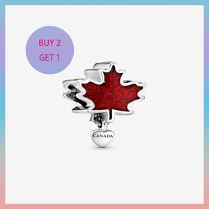 Puede incluir: Un charm de plata con un diseño de hoja de arce roja, un símbolo de Canadá. El charm tiene un colgante en forma de corazón con la inscripción "CANADA". Un círculo morado con el texto "BUY 2 GET 1" está en el fondo.