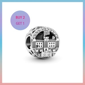 May include: A silver charm bead featuring a detailed depiction of a building with a flag, possibly Buckingham Palace. The charm is adorned with clear crystals and has the word "LONDON" inscribed on the side. A purple circle with the text "BUY 2 GET 1" is in the background.