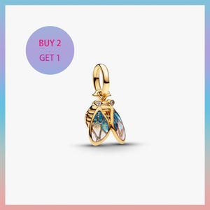 Puede incluir: Un charm dorado con forma de insecto estilizado con alas esmaltadas en azul, blanco y dorado. El charm tiene un lazo para sujetarlo a una pulsera o collar. La imagen incluye un círculo morado con el texto "BUY 2 GET 1".