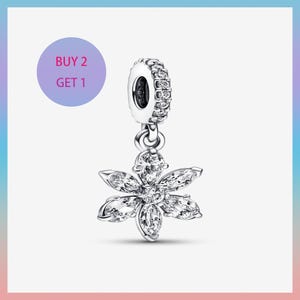 May include: A silver charm with a flower pendant. The flower has six clear, sparkling petals. The charm has a round top with a ring of small, clear stones. A purple circle with the text "BUY 2 GET 1" is in the background.