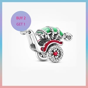 Puede incluir: Un charm de plata con forma de carruaje, con la parte superior verde y detalles rojos. Las ruedas están adornadas con piedras transparentes y un corazón rojo. El charm tiene un asa en forma de corazón. El texto "BUY 2 GET 1" está en un círculo morado.