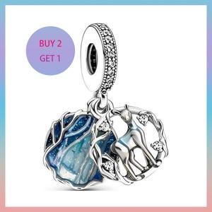 May include: Silver charm featuring a deer design within a circular frame, and a blue and white abstract design. The charm has a decorative top with clear stones. The text "BUY 2 GET 1" is in a purple circle.