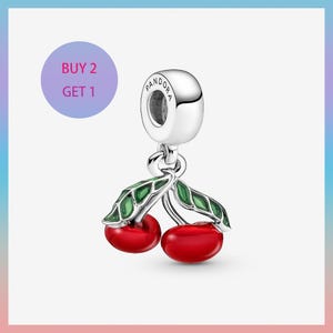 Puede incluir: Un charm de plata con diseño de cereza. El charm presenta dos cerezas rojas brillantes con hojas verdes. El charm tiene un lazo plateado en la parte superior con la marca "Pandora" grabada. Un círculo morado con el texto "BUY 2 GET 1" está en el fondo.