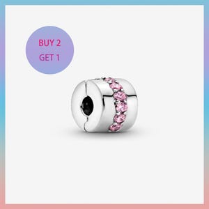 Może przedstawiać: Srebrny charms z rzędem różowych kamieni szlachetnych. Charms jest cylindryczny z czarnym otworem z jednej strony. Fioletowe koło z tekstem "BUY 2 GET 1" znajduje się w tle.