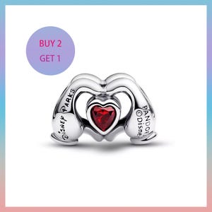 Puede incluir: Un charm de plata con una gema roja en forma de coraz&oacute;n en el centro. El charm tiene la forma de dos manos formando un coraz&oacute;n. Los textos "DISNEY PARKS" y "PANDORA Disney" son visibles. Un c&iacute;rculo morado con el texto "BUY 2 GET 1" est&aacute; en el fondo.
