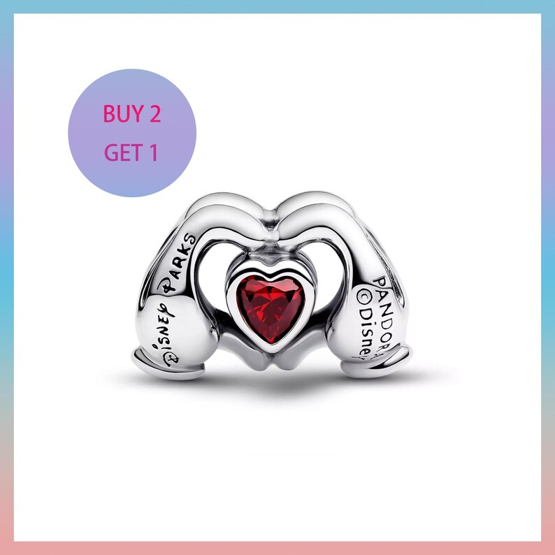 Puede incluir: Un charm de plata con una gema roja en forma de coraz&oacute;n en el centro. El charm tiene la forma de dos manos formando un coraz&oacute;n. Los textos "DISNEY PARKS" y "PANDORA Disney" son visibles. Un c&iacute;rculo morado con el texto "BUY 2 GET 1" est&aacute; en el fondo.