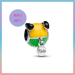May include: A Pandora charm in the shape of a cartoon dog. The charm is yellow, green, and black, with a silver tag that reads "Pluto". The charm has a round shape. The text "BUY 2 GET 1" is in a purple circle.