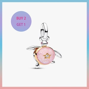 Può includere: Un charm a forma di tartaruga argentato e dorato con un guscio rosa glitterato e un design a forma di stella. Il charm ha una piccola gemma trasparente al centro della stella. L'immagine include anche il testo "BUY 2 GET 1" in un cerchio viola.