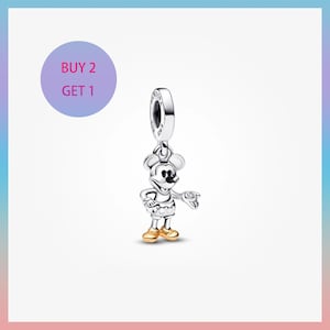 Puede incluir: Un charm de Mickey Mouse plateado con zapatos y un reloj dorados. El charm está suspendido de un lazo plateado. La imagen incluye un círculo morado con el texto "BUY 2 GET 1".