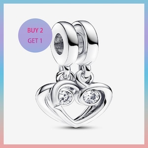 Könnte beinhalten: Ein silberner Charm-Anhänger mit Herzdesign. Der Charm-Anhänger hat zwei klare, runde Edelsteine und den Text "BUY 2 GET 1" in einem lila Kreis. Der Charm-Anhänger ist an zwei kreisförmigen Verbindern aufgehängt.