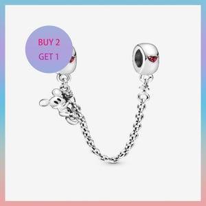 Pode incluir: Uma corrente de segurança de pulseira de berloques prateada com um berloque do Mickey Mouse. A corrente conecta duas contas prateadas, cada uma com um detalhe vermelho em forma de coração. Um círculo roxo com o texto "BUY 2 GET 1" está no fundo.