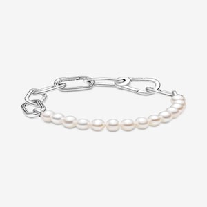 Puede incluir: Una pulsera plateada con un diseño de eslabones de cadena y una hilera de perlas blancas. La pulsera tiene un cierre de broche. Las perlas son pequeñas y están espaciadas uniformemente. La pulsera se muestra sobre un fondo blanco.