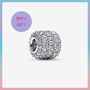 Puede incluir: Una cuenta de dije plateada cubierta de pequeños cristales transparentes. La cuenta tiene forma cuadrada redondeada con un agujero grande en un lado. Un círculo púrpura en el fondo tiene el texto "BUY 2 GET 1".