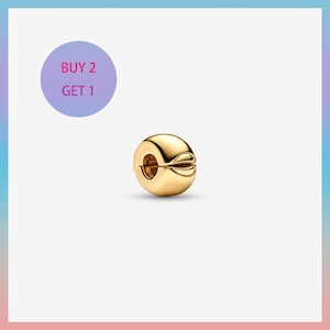 Puede incluir: Un charm dorado con un diseño único. El charm es redondo con un diseño dividido en forma de pétalo en un lado. La imagen también incluye un círculo morado con el texto "BUY 2 GET 1".