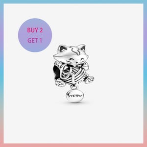 Può includere: Un charm d'argento raffigurante un gatto che tiene una palla di filo. Il charm ha un pendente rotondo con la scritta "MEOW" incisa. Un cerchio viola con il testo "BUY 2 GET 1" è sullo sfondo.