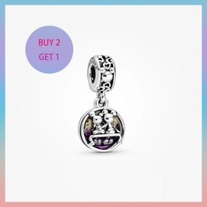 May include: A silver charm with a round pendant featuring two cartoon figures. The charm has a decorative top with heart and Mickey Mouse head cutouts. The pendant has a purple and yellow background. The text "BUY 2 GET 1" is in a purple circle.