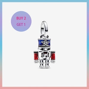 May include: A silver-colored charm in the shape of a toy soldier nutcracker. The charm has a blue and red color scheme with a star on the hat. The text "BUY 2 GET 1" is in a purple circle in the background.