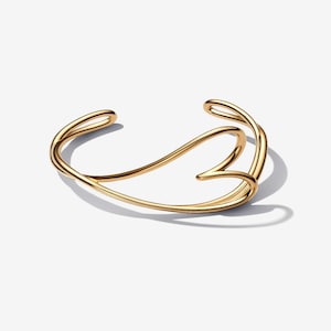 May include: A gold-coloured cuff bracelet with a flowing, open design. The bracelet curves and swirls, creating an elegant, abstract shape. The metal has a polished, reflective surface, and the bracelet is set against a white background.
