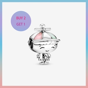 Può includere: Un charm d'argento a forma di personaggio dei cartoni animati sorridente con un cappello con accenti rosa e verdi. Il charm ha un abito dettagliato ed è progettato come ciondolo o perla. Un cerchio viola con il testo "BUY 2 GET 1" è sullo sfondo.