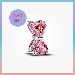 Puede incluir: Un charm de plata con dos cristales rosas en forma de corazón, unidos por una banda plateada. El charm tiene un diseño de lazo. Un círculo morado con el texto "BUY 2 GET 1" está en la esquina superior izquierda.